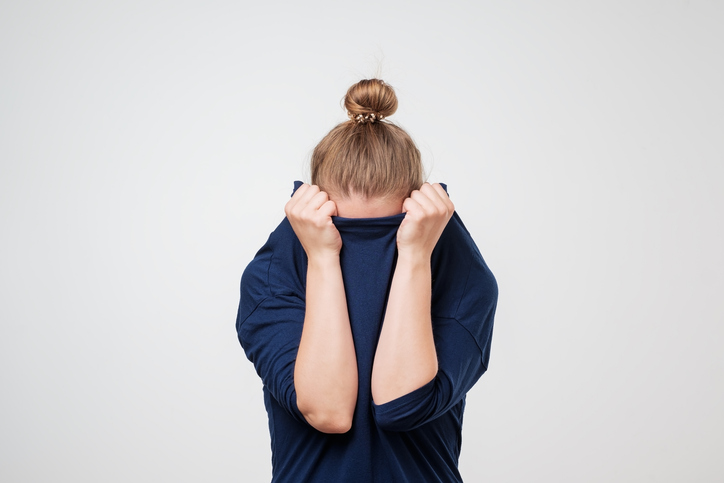 A young woman with a hair bun hides her face in her blue sweater so you can't see her face and is standing against a gray background