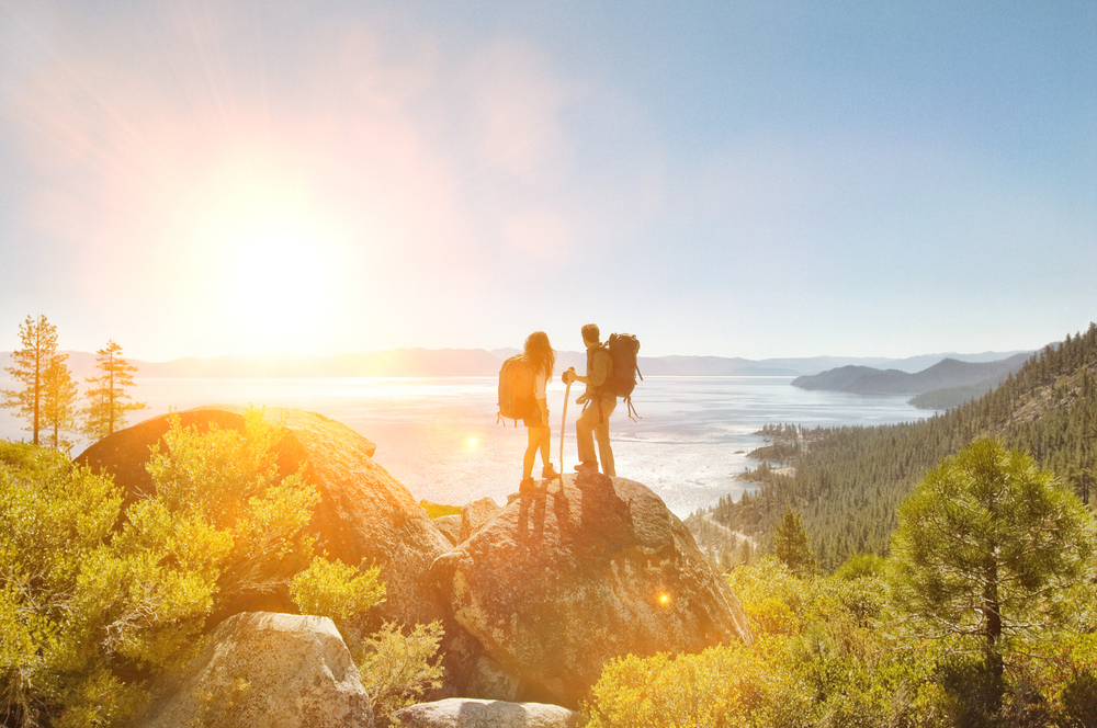 A hiking couple wearing backpacks stands on top of a large rock amid the forest and looks out at the view of the ocean and sunset